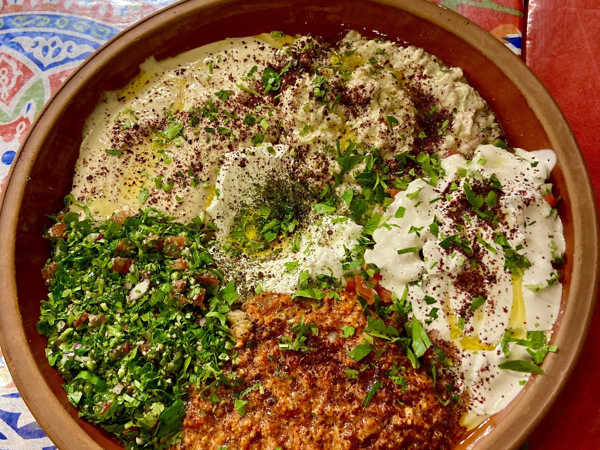 The vibrant mezze platter of hummus, baba ganoush, muhammarah, tahini, tabouleh, labne dusted with sumac is presented in a clay bowl on top of a colorful tablecloth.