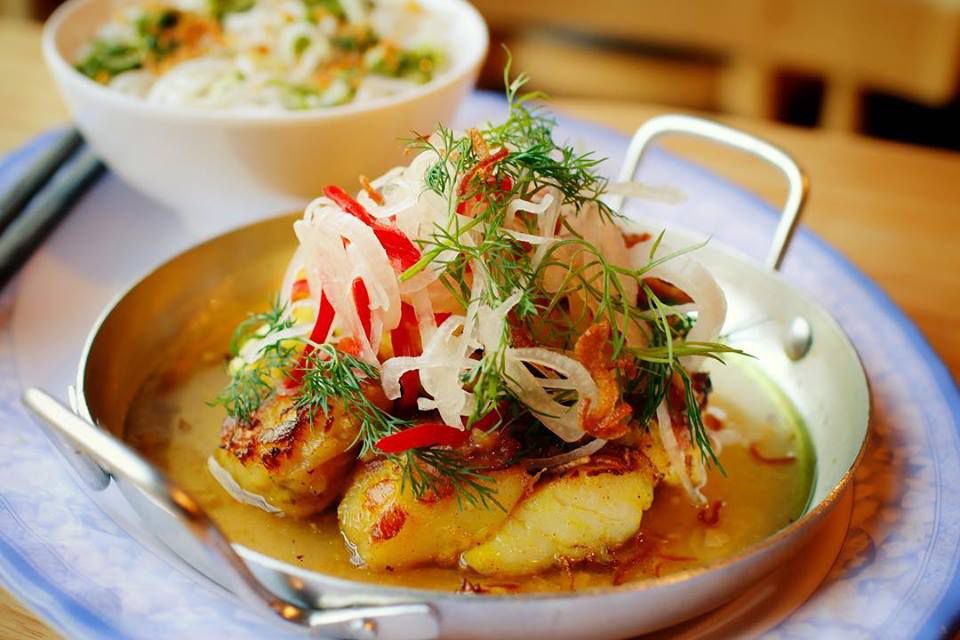 A small metal pan holds a colorful monkfish dish topped with green dill, pickled shallots, and fresno.