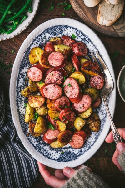 Hands serving a platter of sheet pan sausage and potatoes