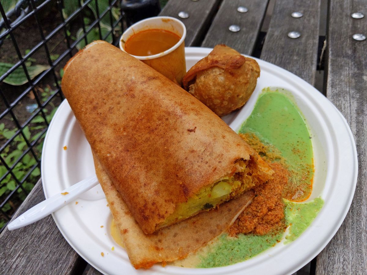 The image showcases a white paper plate containing a dosa, a green cilantro sauce, a samosa, and a red sauce in a plastic cup, all placed on a wooden bench.