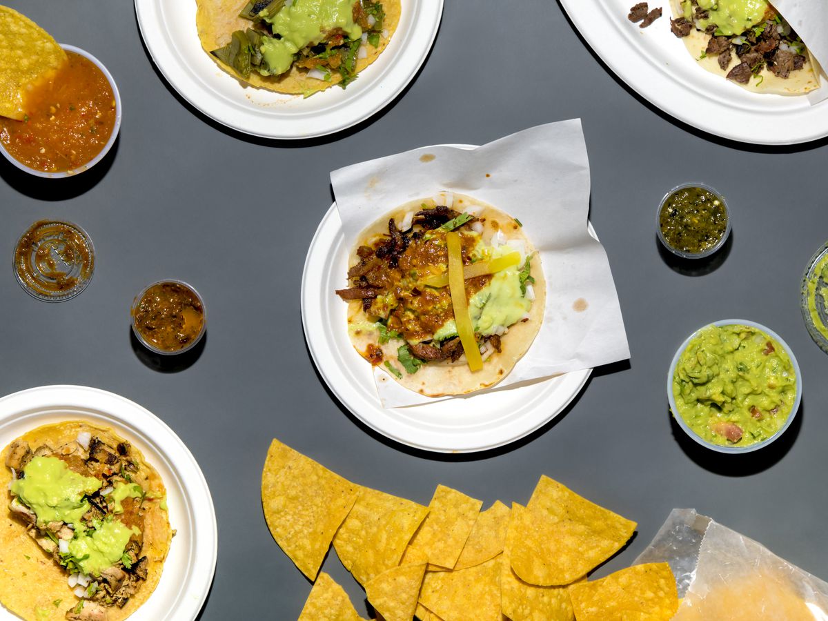 The image shows an overhead view of tacos, chips, guacamole, and plastic containers of salsa.